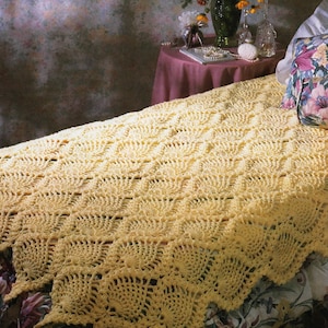 May include: A handmade, yellow crochet blanket with a scalloped edge is draped over a bed. The blanket's intricate pattern creates a textured surface. The bed has a floral patterned bedspread and a pillow with a floral design.