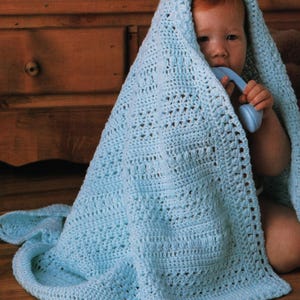 May include: A baby wrapped in a light blue crocheted blanket. The blanket has a textured pattern and is draped over the baby's head. The baby is holding a blue toy. The blanket is on a wooden surface.