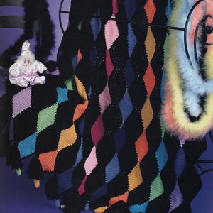 May include: A black knitted blanket with a diamond pattern in various colors, including pink, orange, green, and blue. The blanket is draped over a black metal rack. A doll and a feather boa are also visible.