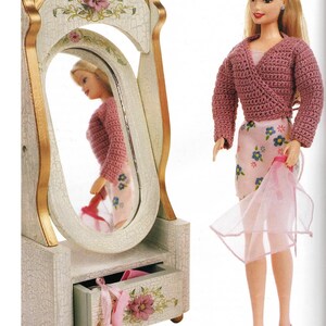 Puede incluir: Una muñeca con un suéter envolvente de ganchillo rosa y una falda floral, sosteniendo una tela rosa transparente. Un espejo de tocador decorativo con un cajón está a la izquierda, con detalles florales y zapatillas de ballet rosas.