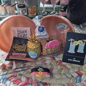 May include: A MGA's Miniverse Make It Mini Happy Hour kit featuring a gold pineapple-shaped cup, a bag of pebble ice, a small box with pink coasters, a pair of tweezers, and a recipe card for The Blue Hawaiian cocktail with two blue drinks.