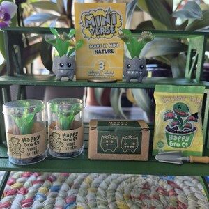 May include: A MGA's Mini Verse Make It Mini Nature Series 3 kit featuring two small grey monster-faced planters with green Venus fly trap plants, two clear containers with plant labels, a brown box, a green seed packet, and a small metal shovel.