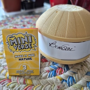 May include: A yellow and white Mini Verse box with the text "Make it Mini Nature" and a number 3. Next to it is a tan and white container with the word "Bonsai" written on it. The items are on a colorful braided mat.