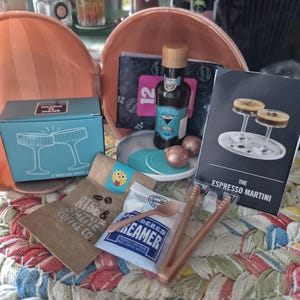 May include: A miniature espresso martini cocktail kit featuring a small bottle labeled Espresso Martini, a blue box with two coupe glasses, a Rise & Shine Coffee Co. bag, a powdered creamer packet, a small spoon, tongs, and a recipe card for an espresso martini.