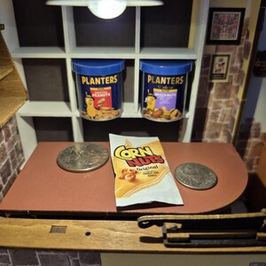May include: Miniature scene with two Planters peanut jars and a bag of Corn Nuts on a red countertop. Two coins are on the countertop. The scene is lit by a small overhead light.