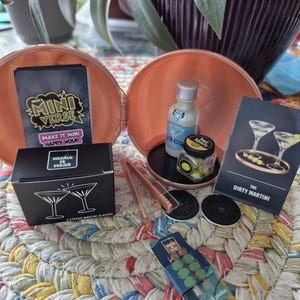 May include: A Mini Verse Make It Mini Happy Hour kit featuring a black box labeled 2 Etched Martini Glasses, a small bottle of liquid, a jar of green olives, a pair of copper-colored tongs, two black coasters, and a Dirty Martini recipe card.