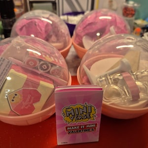 May include: Several Mini Verse Valentine's themed surprise balls with clear plastic domes and pink bases. One box is visible, featuring the text "Mini Verse" and "Make it Mini Valentine's". The balls contain miniature items and pink tissue paper.