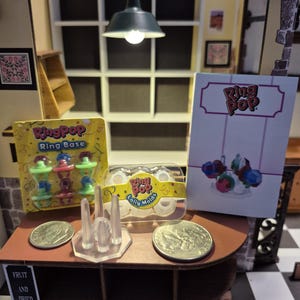 May include: A miniature candy shop display featuring Ring Pop products. The display includes Ring Pop ring bases, lolly molds, and promotional materials. The shop has a brown countertop, shelves, and a sign that reads "Chocolate Workshop".
