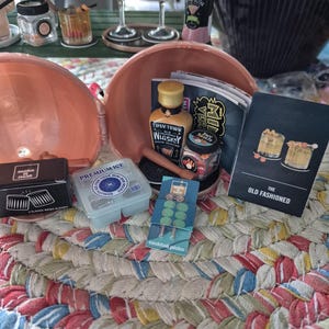 May include: A miniature bar set featuring a bottle of Tipsy Town whiskey, a container of premium ice, a box of two classic rocks glasses, a pack of cocktail picks, and a recipe card for an Old Fashioned, all housed in a peach-colored container.