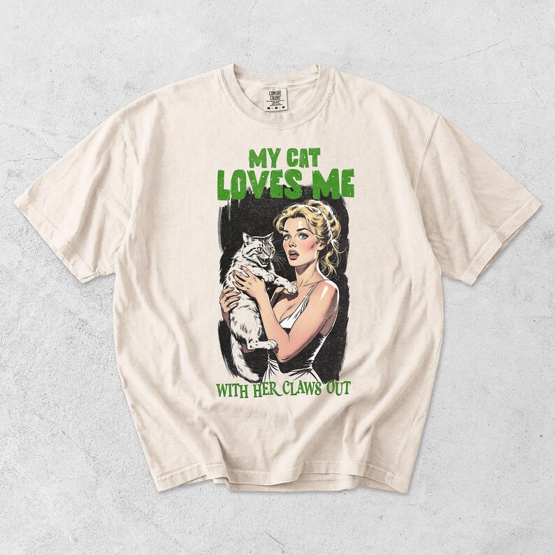 K&ouml;nnte beinhalten: Ein cremefarbenes Kurzarm-T-Shirt mit einer grafischen Illustration einer Person, die eine Katze h&auml;lt. Der Text MY CAT LOVES ME steht oben und WITH HER CLAWS OUT unten in gr&uuml;ner Schrift. Die Grafik hat einen schwarzen Hintergrund.