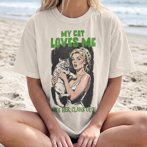 K&ouml;nnte beinhalten: Ein cremefarbenes T-Shirt mit einer grafischen Illustration einer Person, die eine Katze h&auml;lt. Der Text lautet MY CAT LOVES ME WITH HER CLAWS OUT in gr&uuml;ner Schrift. Die Illustration zeigt eine Person mit blonden Haaren und eine Katze mit sichtbaren Krallen.