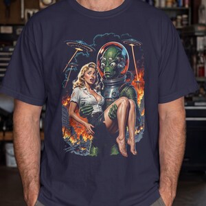 Può includere: Una t-shirt blu scuro con un'illustrazione grafica di un alieno verde multi-occhi in tuta spaziale che trasporta una persona bionda con camicia bianca e gonna scura. Lo sfondo raffigura una città in fiamme con dischi volanti nel cielo.