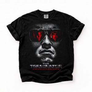 Könnte beinhalten: Ein schwarzes Kurzarm-T-Shirt mit einer Grafik eines Gesichts, das eine Sonnenbrille trägt, in der sich zwei Personen spiegeln. Unter dem Gesicht steht der Text THE TERMINATOR sowie THE THING THAT WON'T DIE, IN THE NIGHTMARE THAT WON'T END in Weiß und Rot.