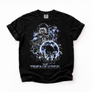 Könnte beinhalten: Ein schwarzes T-Shirt mit einem Grafikdruck eines metallischen Roboterschädels mit leuchtend roten Augen, einer Silhouette in einer blauen Kugel und Blitzeffekten. Der Text THE TERMINATOR ist in einer metallischen Schriftart unten aufgedruckt.