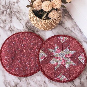 May include: Two round, quilted fabric trivets with a floral pattern and a starburst design. The trivets are burgundy with red trim. A small woven basket with beige pom-pom flowers sits in the background.