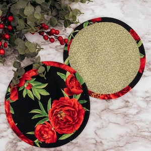 May include: Two round coasters. One coaster features a black background with red floral designs and green leaves. The other coaster has a tan fabric center with a red and black floral border. Decorative greenery and berries are in the background.