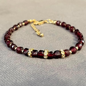Puede incluir: Una pulsera de cuentas con piedras preciosas facetadas de color rojo intenso y detalles dorados. La pulsera tiene una cadena y un cierre dorados. Los detalles dorados incluyen pequeños elementos decorativos con piedras claras y brillantes.
