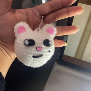 May include: A white crocheted keychain in the shape of a bear's head. The bear has pink ears, black eyes, a pink nose, and a small black dot. The keychain is attached to a silver metal ring.
