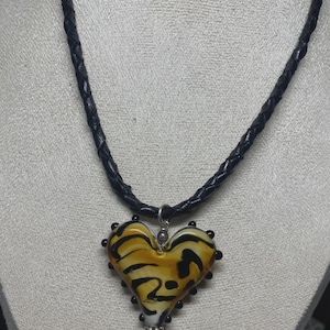 May include: A heart-shaped pendant necklace with a black braided leather cord. The pendant is glass, featuring a yellow and black tiger stripe design, and is accented with small black beads around the edges.