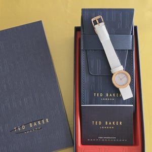 May include: A Ted Baker London watch with a white strap and gold-tone case, presented in a navy blue box with the brand's logo. The watch is displayed in a navy blue case with a red interior, alongside a user information card.