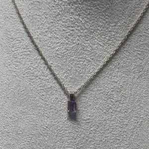 May include: A silver necklace with a rectangular, light purple gemstone pendant. The pendant is suspended from a delicate chain. The necklace is displayed against a textured, light gray background.