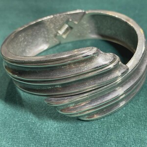May include: Silver-toned hinged bracelet with a ridged design. The bracelet has a clasp closure and a curved, wave-like pattern on the front. The jewelry is displayed on a teal green surface.