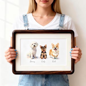 May include: A framed horizontal print featuring a watercolor-style illustration of three dogs sitting side-by-side. From left to right, the dogs are a white Bichon Frise named Benny, a small black and tan terrier named Cody, and a tan and white Corgi named Rick.