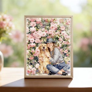 Pode incluir: Uma impressão em aquarela emoldurada com uma mulher e um cão golden retriever em frente a um fundo de rosas cor-de-rosa. A mulher usa uma camisa listrada, jeans e um chapéu. A moldura é de cor castanho claro.