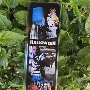 Puede incluir: Un marcapáginas rectangular con temática de Halloween, que muestra imágenes de la película. La palabra "HALLOWEEN" está impresa en letras blancas en negrita. Las imágenes incluyen una lápida, una máscara de payaso y una calabaza tallada.