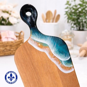 May include: Wooden chopping board with a handle, featuring a resin design. The handle is black, transitioning to a blue and white wave pattern. The board has a natural wood grain finish and a circular hole in the handle. The text "FAIT AU QUEBEC" and "MADE IN QUEBEC" is visible.