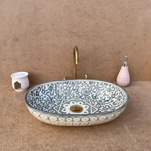 Puede incluir: Lavabo ovalado de cerámica con un detallado estampado floral azul y blanco. El lavabo tiene un grifo dorado y está acompañado de un pequeño recipiente de cerámica blanca y un dispensador de jabón rosa. Un accesorio de baño decorativo.