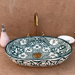 May include: Oval ceramic sink with a teal and white floral pattern. The sink has a gold-colored faucet and drain. A pink soap dispenser and a small white dish are next to the sink.