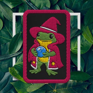 May include: Embroidered patch showing a green frog in a red wizard hat and cloak, holding a blue crystal ball. The patch has a black background with a red border and white details. The frog has yellow accents.