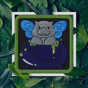 Può includere: Toppa ricamata con un ippopotamo grigio con ali di fata blu seduto in un calderone blu scuro con melma verde. La toppa ha uno sfondo nero con un bordo verde, su un quadrato bianco.