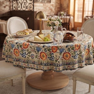 May include: A round wooden dining table covered with a cream-colored tablecloth featuring a vibrant floral pattern of orange, red, and blue flowers. The table is set with a glass pitcher of water, a plate of cheese, grapes, and bread.
