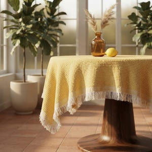May include: A round yellow tablecloth with a waffle-knit texture and white fringe trim draped over a pedestal table. On the table sits a small amber glass vase containing dried pampas grass stems and a single yellow lemon beside the vase.