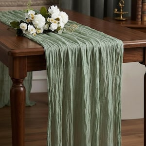 Può includere: Runner da tavolo strutturato verde salvia su un tavolo in legno scuro. Un bouquet di fiori bianchi con steli e foglie verdi poggia sul runner. Il runner ha un aspetto increspato e fluido, aggiungendo un tocco di eleganza.
