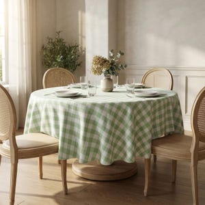 May include: A round dining table covered with a light green and white gingham checkered tablecloth. The table is set with four white plates, four clear drinking glasses, and a central ceramic vase containing a bouquet of dried flowers.