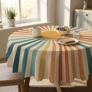 Puede incluir: Un mantel redondo con un patrón de rayos de sol en tonos naranja, amarillo y azul verdoso sobre un fondo crema. La tela tiene un borde festoneado y cubre una mesa con una taza de cerámica, un cuenco de fruta y una rebanada de pan.