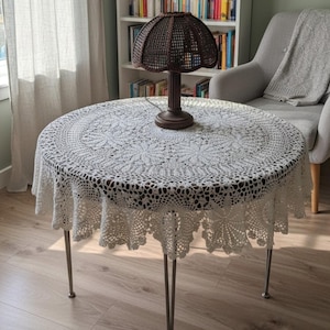 Può includere: Un tavolo rotondo con sottili gambe in metallo è coperto da una tovaglia circolare in uncinetto bianco con un intricato motivo floreale e un bordo smerlato. Una lampada da tavolo marrone scuro con paralume intrecciato è al centro.