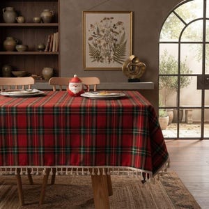 May include: A rectangular tablecloth featuring a red, green, and white plaid pattern with a cream-coloured fringe trim along the edges. The fabric is draped over a wooden table, creating a festive appearance for a dining room setting.