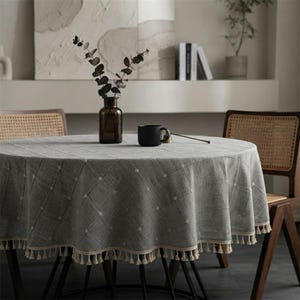 May include: A round grey tablecloth with a subtle white grid pattern and a cream-coloured fringe trim along the edge. The tablecloth is draped over a round table, featuring a dark brown glass vase with dried branches and a black ceramic mug.