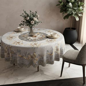 May include: A round tablecloth featuring a light gray and white floral pattern with gold accents. The design includes a central circular motif, a repeating flower border, and flowing leaf details. A glass vase with flowers and two bowls sit on top.