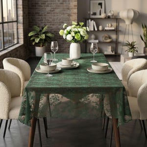 May include: A rectangular dining table covered with a dark green lace tablecloth featuring a floral pattern. The table is set with four white ceramic bowls on matching plates, four clear wine glasses, and a white vase filled with white flowers.