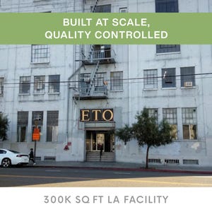 May include: Exterior view of a large, light-colored building with multiple windows and a fire escape. The text "BUILT AT SCALE, QUALITY CONTROLLED" is displayed across the top. The building entrance has the letters "ETO" above it. The text "300K SQ FT LA FACILITY" is at the bottom.