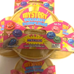 May include: A collection of Mystery Squashy Dumplings toy sets in round yellow containers. The pink packaging features the text Mystery Squashy Dumplings, Metallic, and age 5+. Each pack includes one dumpling and a steamer pot, with illustrations of various colored dumplings with faces.
