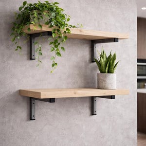 Op de afbeelding: Twee zwevende houten planken met zwarte metalen beugels aan een grijze muur. De bovenste plank heeft een geweven mand met hangende groene planten. De onderste plank toont een pot met slangengras in een betonnen pot. Woondecoratie.