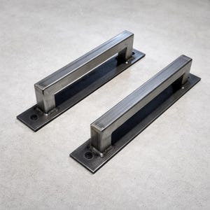 Może przedstawiać: Dwa uchwyty do drzwi w stylu industrialnym. Każdy uchwyt ma prostokątną metalową podstawę ze spawanym, prostokątnym uchwytem. Metal ma szczotkowane, srebrne wykończenie. Podstawa jest mocowana czarnymi śrubami.