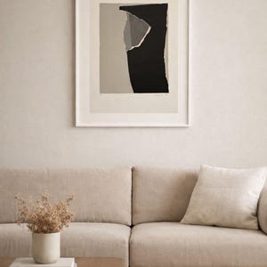 May include: Abstract art print in a white frame. The artwork features a torn paper design with black, gray, and beige shapes. The print hangs above a beige sofa with a matching pillow. A wooden coffee table and a vase with dried flowers are in front of the sofa.