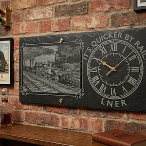 Op de afbeelding: Een zwarte leien klok met een treinillustratie en de tekst "It's Quicker By Rail" en "LNER". De klok heeft gouden wijzers en Romeinse cijfers. Een ingelijste treinposter is ook zichtbaar.
